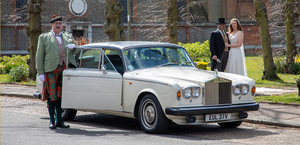Jonathan Sayers of Concierge Wedding Cars standing by his 1980 Rolls Royce Silver Wraith MKII; Bride and Groom stand together in the background.